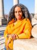 Portrait of Alyscia Cunningham, Trinidadian-American artist. Her hair is out in a crinkled look and she has a mustard yellow knitted Pancho. She rests her elbow on the base of the column beside her. Two other columns can be seen in the background with the blue sky in-between. .